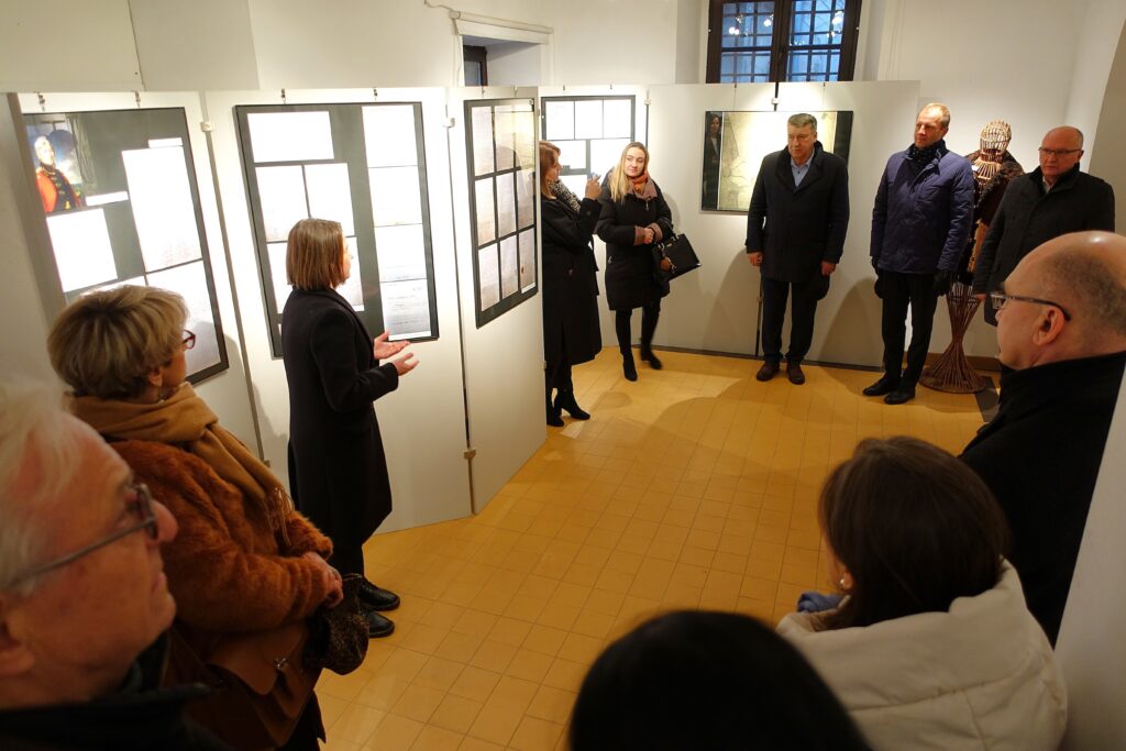 Grupa osób w ciemnych płaszczach stoi w galerii z białymi ścianami i żółtą podłogą, oglądając wystawę dokumentów.