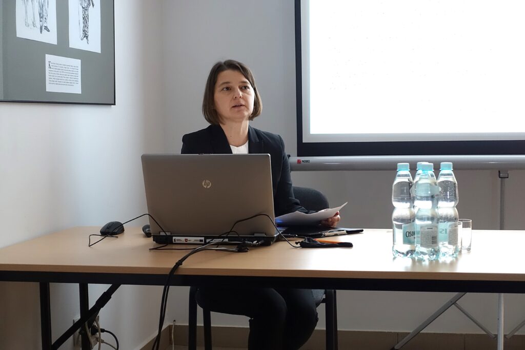 Kobieta w czarnym garniturze siedzi przy stole z laptopem, trzema butelkami wody i szklanką, obok jasny ekran projekcyjny.