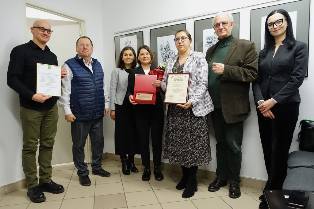 Siedem osób stoi na jasnej ścianie z czarno-białymi rysunkami, dwie trzymają dyplomy i kwiaty, ubrani formalnie i półformalnie.