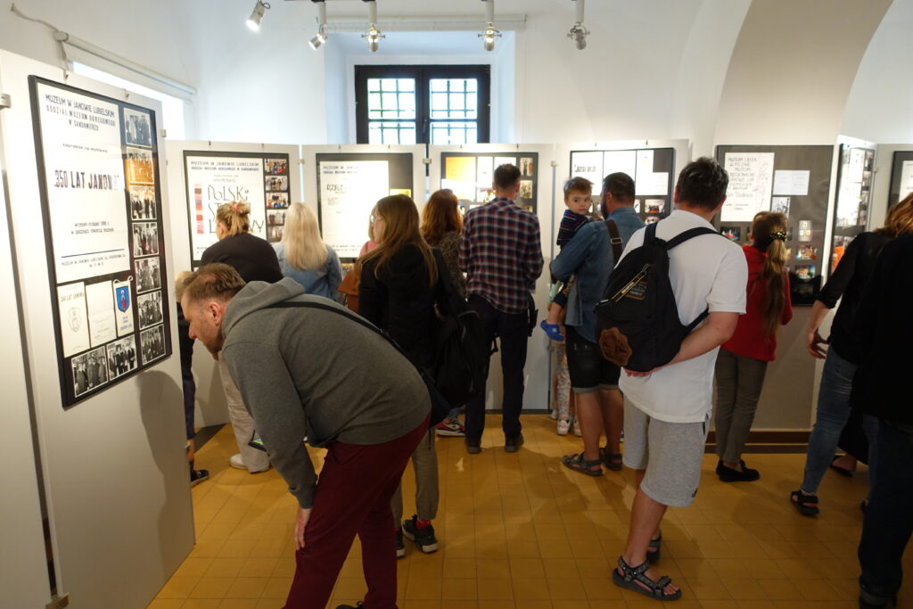 Ludzie oglądają wystawę w jasnym pomieszczeniu z tablicami informacyjnymi i oknem z kratami.