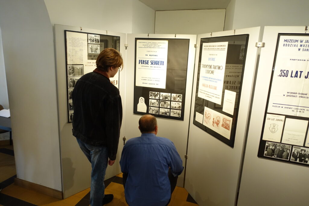 Dwóch mężczyzn ogląda plansze z tekstami i czarno-białymi zdjęciami na białych ściankach wystawowych w pomieszczeniu.