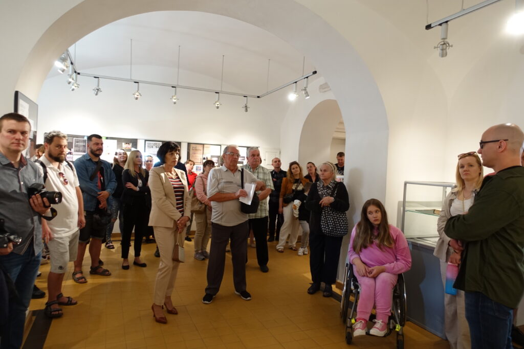 Grupa ludzi w jasnym pomieszczeniu z łukowatym sufitem, niektórzy stoją, a jedna dziewczyna siedzi na wózku inwalidzkim.