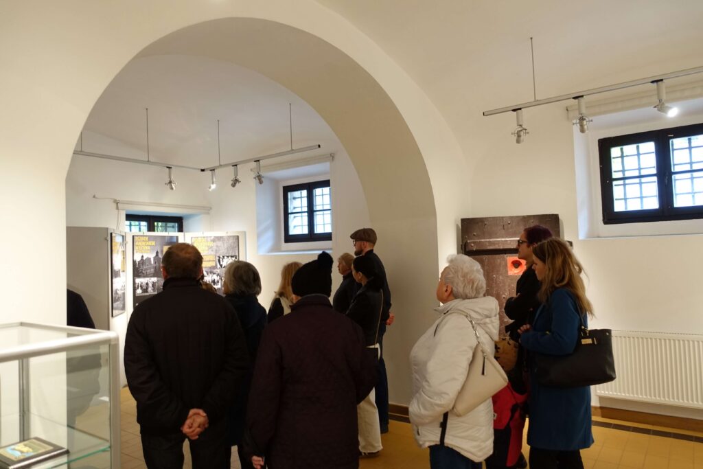Grupa osób w różnym wieku stoi w jasnym pomieszczeniu z łukowym stropem, oglądając wystawę na ścianie.