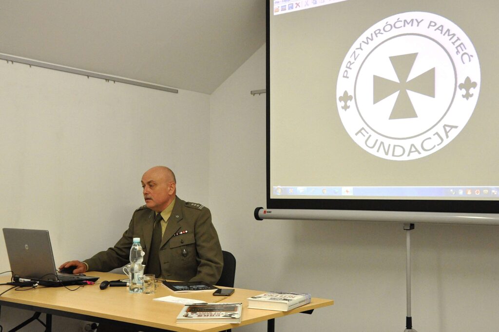 Mężczyzna w mundurze siedzi przy stole z laptopem, butelką wody i książkami, obok biały ekran z logo fundacji.