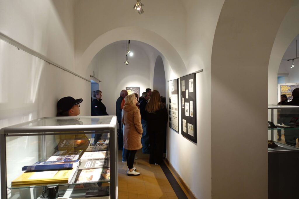 Grupa osób oglądająca wystawę na białej ścianie w pomieszczeniu z łukowatym stropem, obok szklana witryna z książkami.