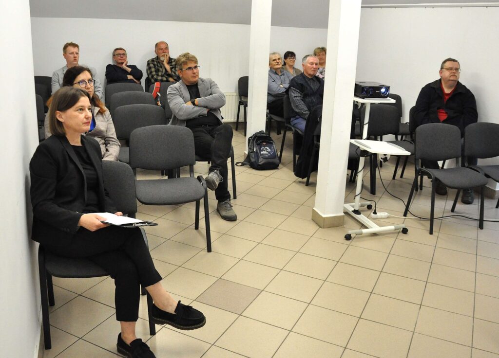 Grupa osób siedzi na szarych krzesłach w jasnym pomieszczeniu z białymi ścianami i jasnobeżową podłogą z płytek ceramicznych.