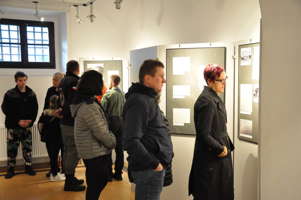 Grupa ludzi w różnorodnych ubraniach stoi i ogląda plansze z tekstem i zdjęciami na jasnych ścianach galerii.