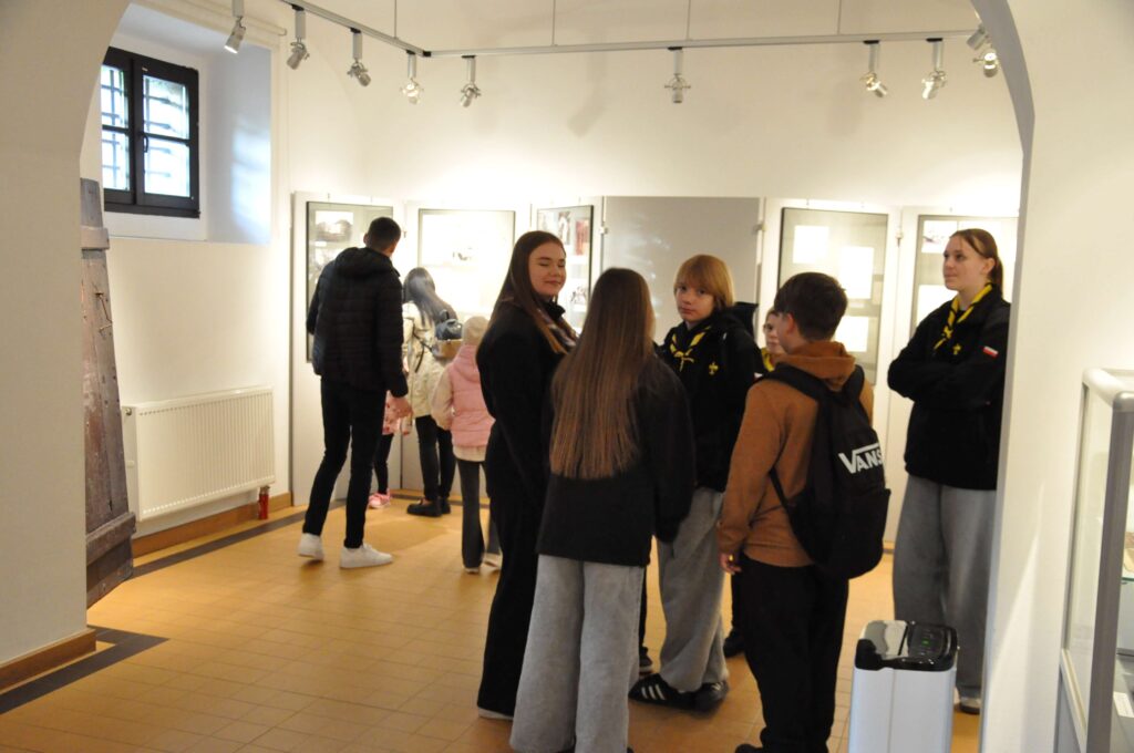 Grupa młodzieży w jasnym wnętrzu galerii z białymi ścianami i tablicami na ekspozycje, część osób ubrana w czarne bluzy.