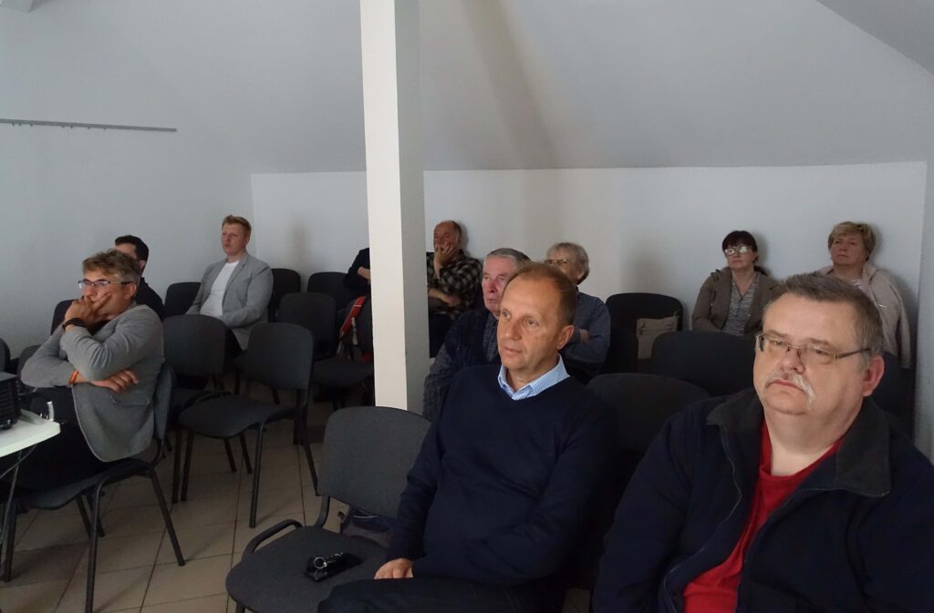 Grupa dorosłych osób siedzi na ciemnoszarych krzesłach w jasnym pomieszczeniu z białymi ścianami i jasnoszarym sufitem.