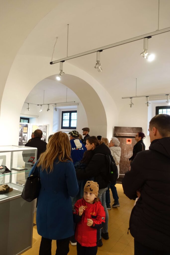 Grupa ludzi w jasnym pomieszczeniu z białymi ścianami i łukowatym stropem, część zwraca uwagę na ekspozycję muzealną.