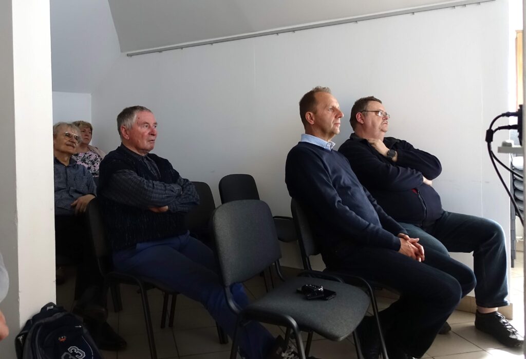 Grupa dorosłych mężczyzn i kobiet siedzi na szarych krzesłach w jasnym, pustym pomieszczeniu.