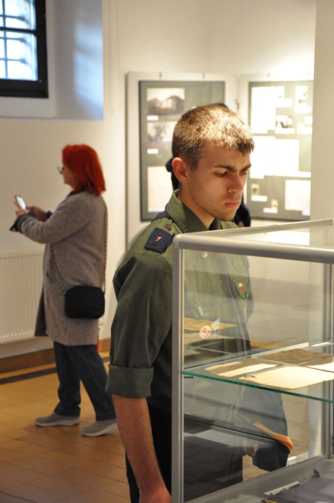 Młody mężczyzna w zielonej koszuli wojskowej ogląda eksponaty w szklanej gablocie, w tle kobieta z czerwonymi włosami korzysta z telefonu.