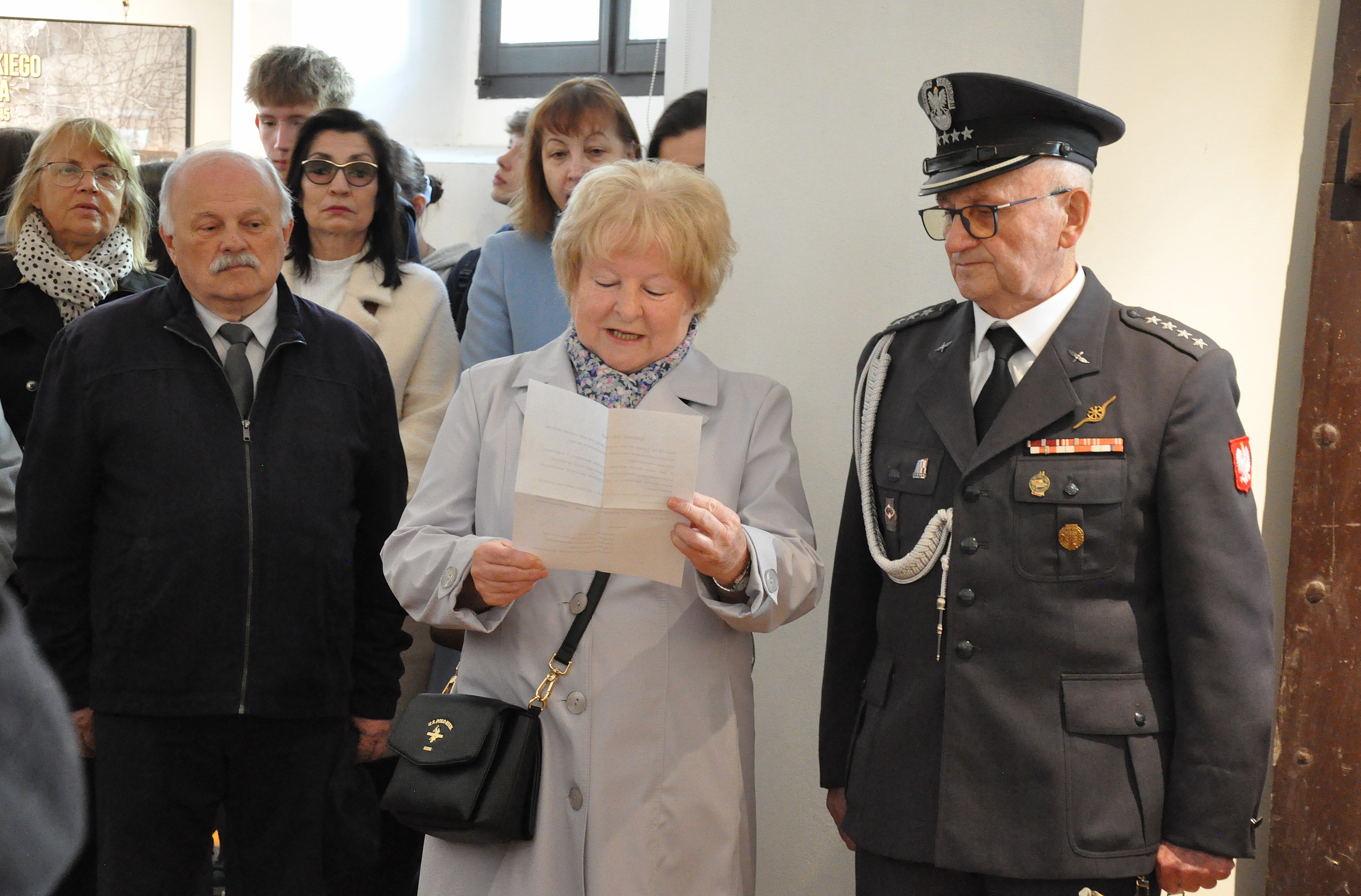 Starsza kobieta czyta dokument, obok mężczyzna w mundurze wojskowym, w tle grupa ludzi, jasne wnętrze z oknem.