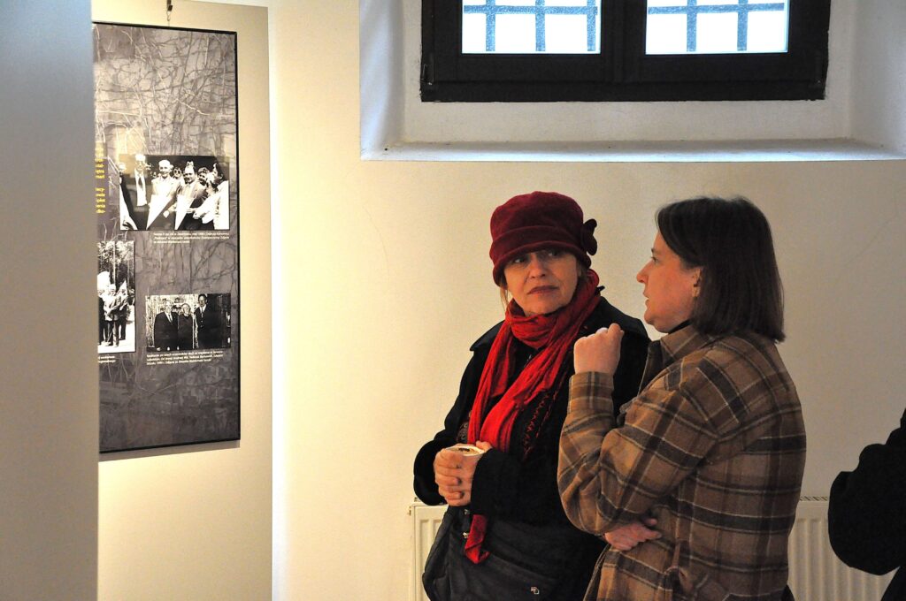 Dwie kobiety rozmawiające w jasnym pomieszczeniu, jedna w bordowym kapeluszu i szalu, druga w brązowej kratowanej kurtce.