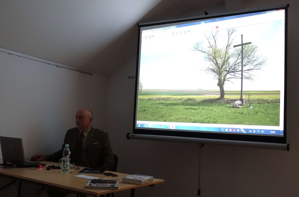 Mężczyzna w mundurze przy stole z laptopem, wodą, książkami i notatnikiem, obok ekran z projekcją pola z drzewem i krzyżem.