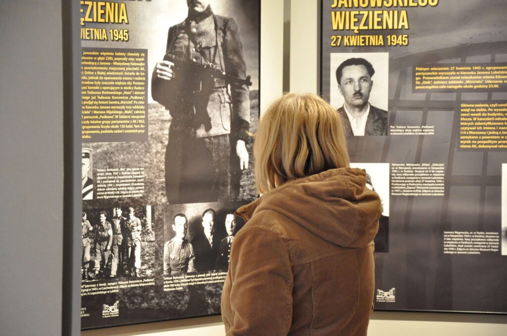 Kobieta w beżowej kurtce stoi tyłem przed tablicami z czarno-żółtymi historycznymi tekstami i zdjęciami mężczyzn.