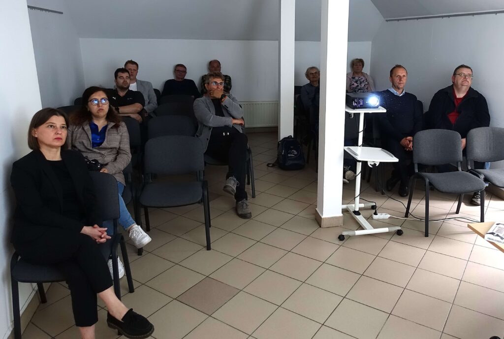 Grupa osób siedzi na krzesłach w jasnym pomieszczeniu z białymi ścianami i beżową podłogą, obok stoi biały rzutnik na statywie.