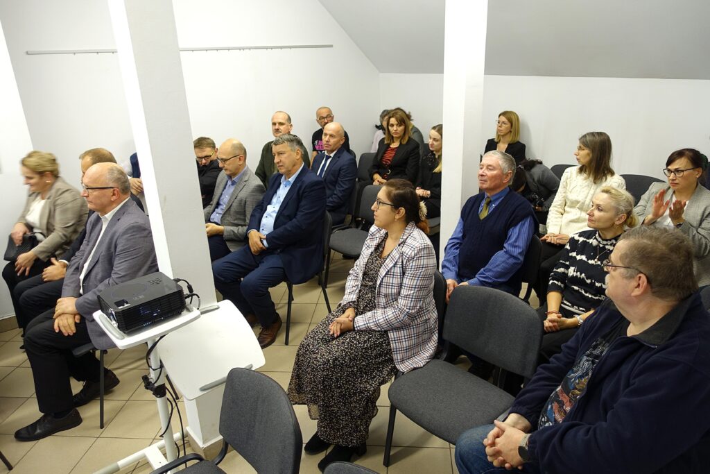 Grupa osób siedzi na krzesłach w jasnym pomieszczeniu z białymi ścianami i dwoma białymi filarami.