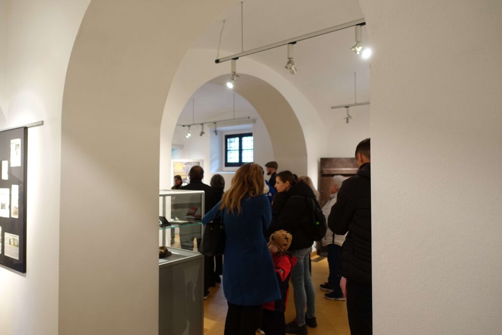 Grupa ludzi w pomieszczeniu z białymi ścianami i łukowymi przejściami, z ekspozycją muzealną i oświetleniem sufitowym.