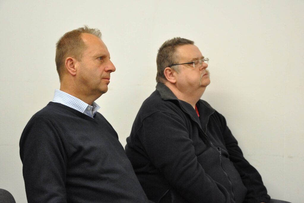 Dwaj mężczyźni w ciemnych ubraniach siedzą na jasnym tle, patrząc w prawo.