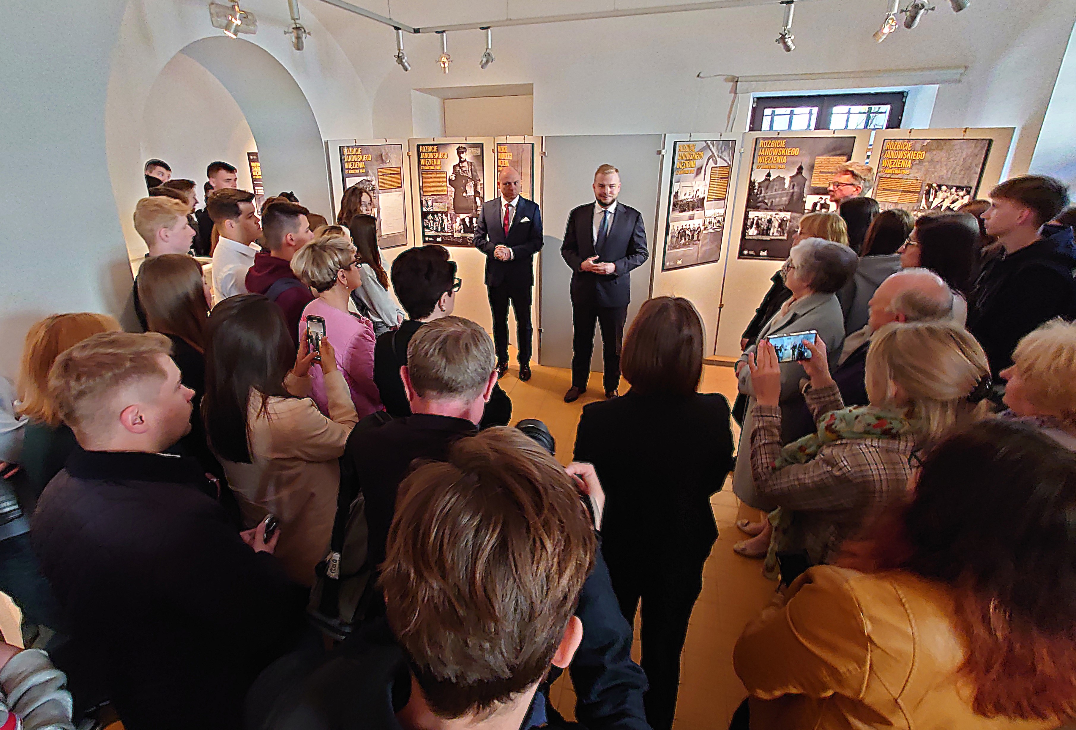 Grupa osób słuchających dwóch mężczyzn w garniturach na tle plansz wystawy w jasnym pomieszczeniu z łukowatym sufitem.