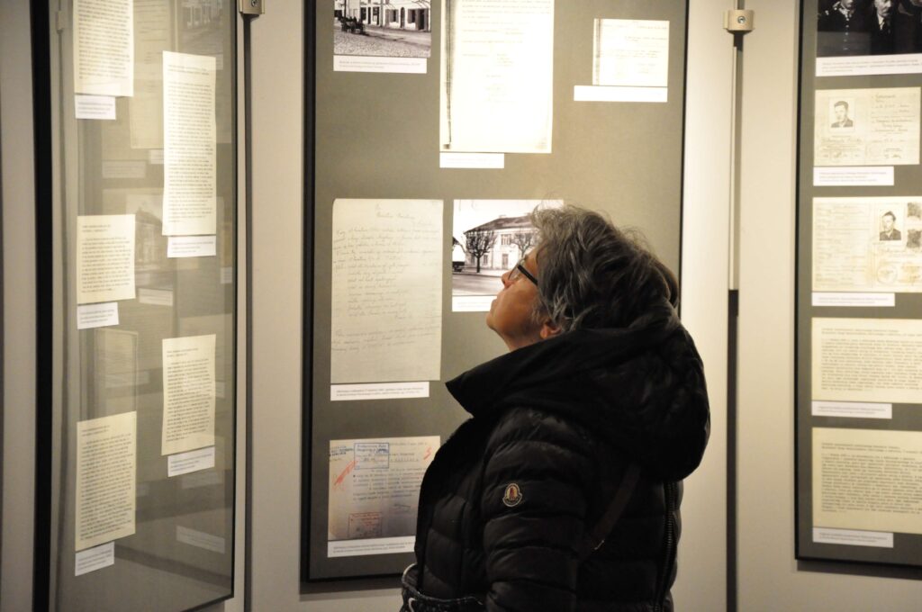 Kobieta w czarnej kurtce z kapturem ogląda dokumenty i zdjęcia w gablotach na jasnej ścianie muzeum.