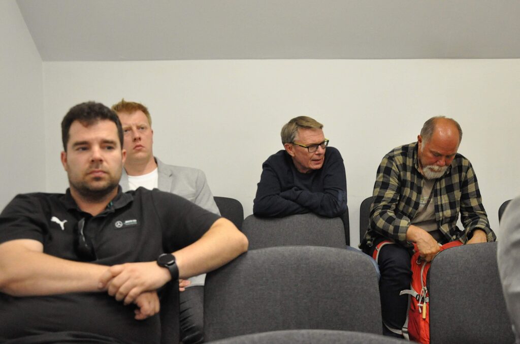 Czterech mężczyzn siedzi na krzesłach, biały sufit i ściana, mężczyzna z prawej szuka czegoś w czerwonym plecaku.