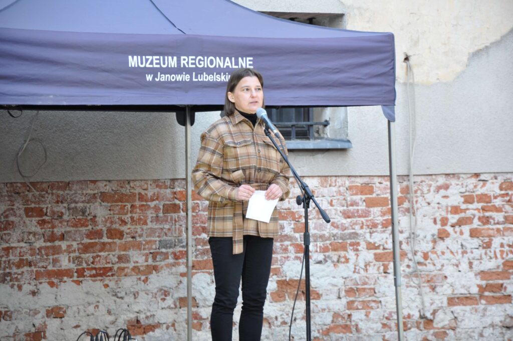 Kobieta w brązowej kraciastej kurtce mówi do mikrofonu przed namiotem z napisem Muzeum Regionalne w Janowie Lubelskim, tło ceglana ściana.