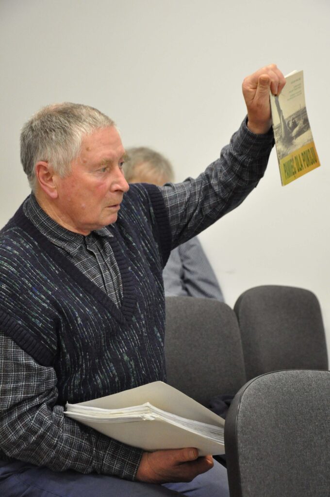 Starszy mężczyzna w ciemnej kamizelce trzyma kartkę z tekstem, siedzi na szarym krześle na jasnym tle.