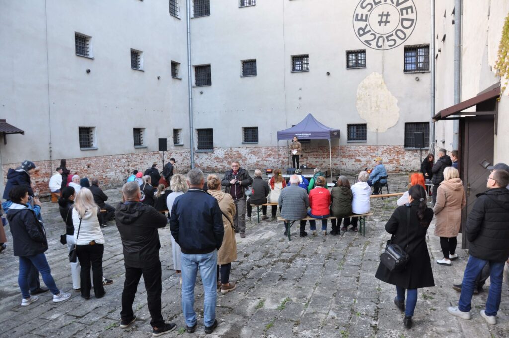 Grupa ludzi na dziedzińcu z kamienną nawierzchnią, siedzących i stojących przed namiotem z mikrofonem.