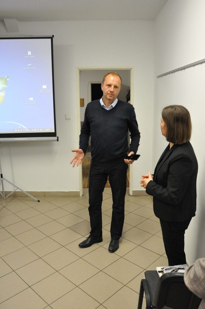 Mężczyzna w ciemnym swetrze i spodniach oraz kobieta w czarnym garniturze stoją w jasnym pomieszczeniu z rzutnikiem i drzwiami w tle.