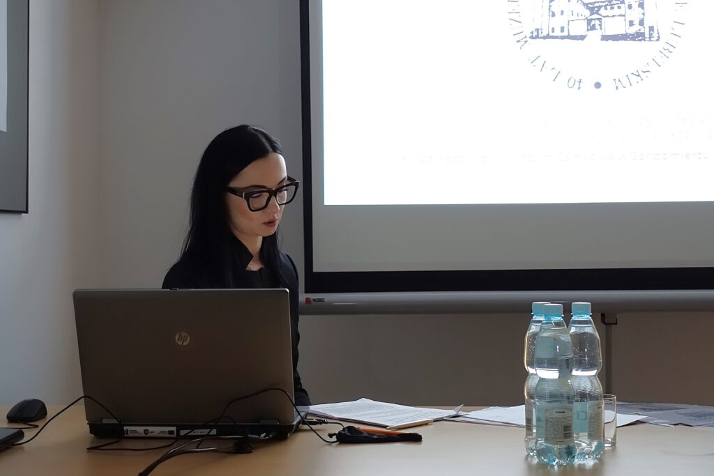 Kobieta z ciemnymi włosami i czarnymi okularami siedzi przy stole z laptopem, dokumentami i butelkami wody, na tle ekranu projekcyjnego.