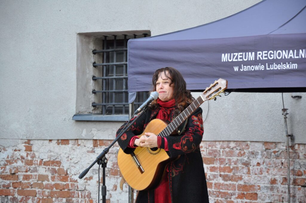 Kobieta w czarnym płaszczu z czerwonymi wzorami gra na gitarze klasycznej przed ścianą z cegły i oknem z kratami.