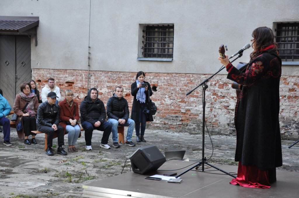 Kobieta w czarno-czerwonym płaszczu gra na gitarze i śpiewa do mikrofonu, ludzie siedzą na ławkach przed nią.