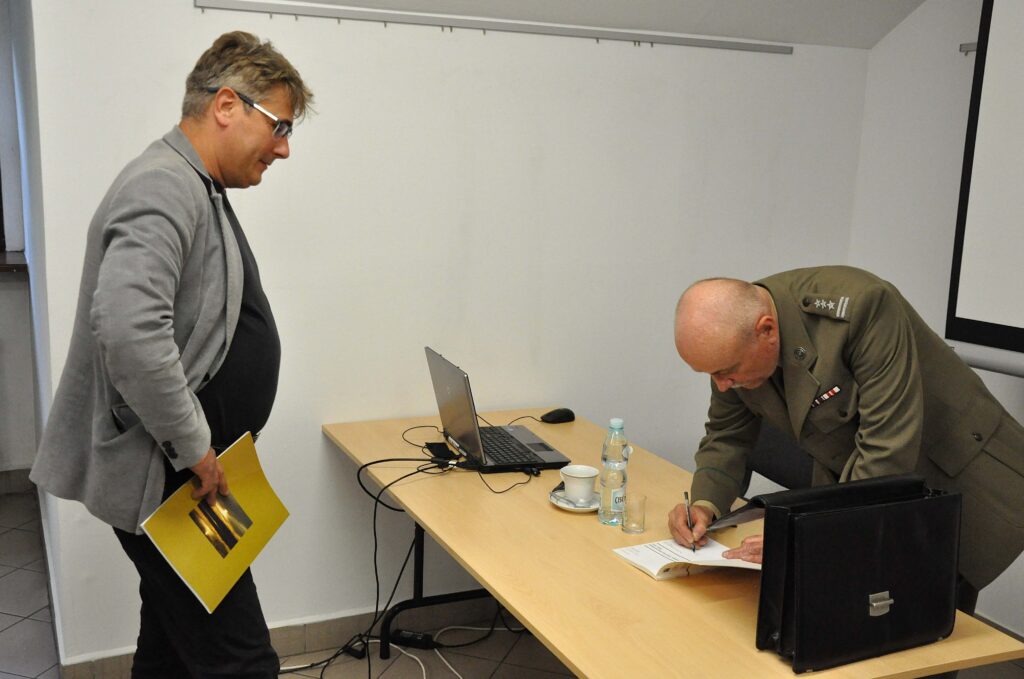 Dwóch mężczyzn przy stole, jeden w garniturze podpisuje książkę, drugi z okularami trzyma żółtą teczkę, na stole laptop, butelka, filiżanka.