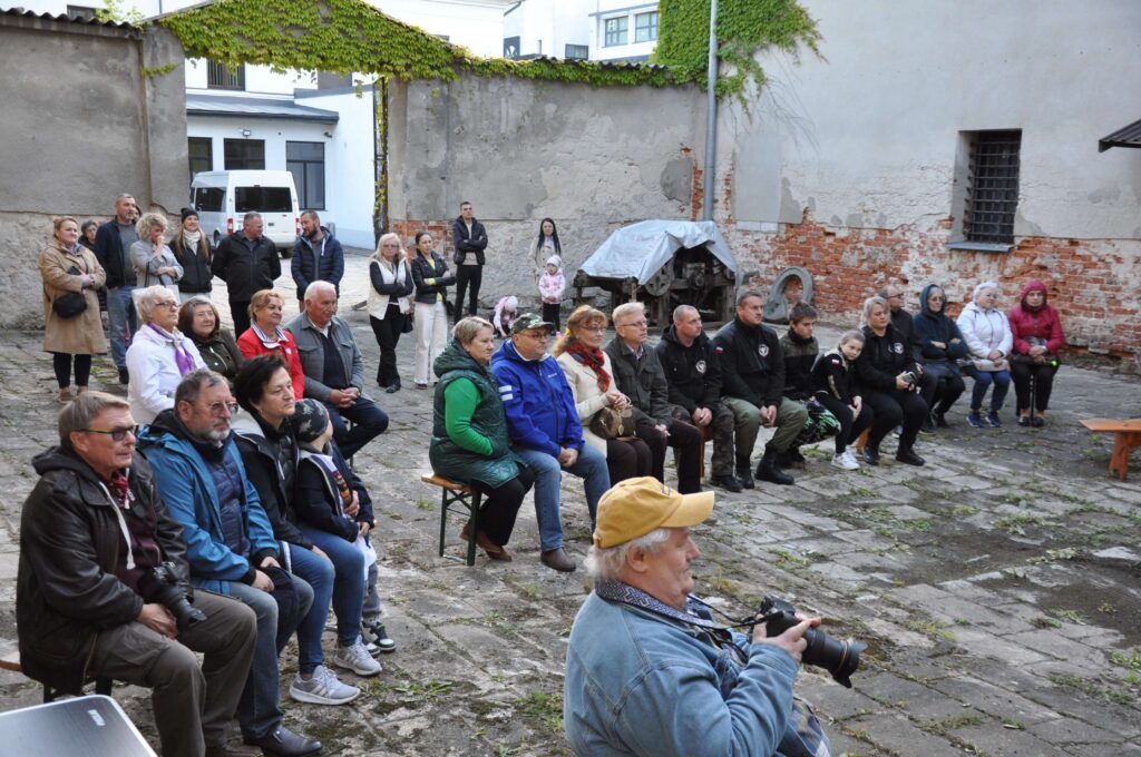 Grupa osób siedzących i stojących na dziedzińcu z kamienną posadzką, stare mury z cegły i bluszczem w tle.