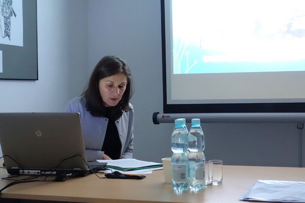 Kobieta w szarej marynarce czyta dokumenty przy stole z laptopem, butelkami wody i szklankami, obok ekran projekcyjny.