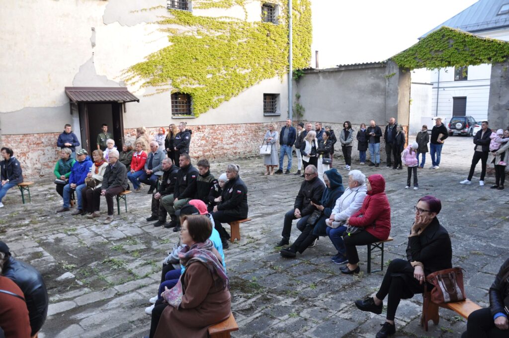 Ludzie siedzący i stojący na kamiennym dziedzińcu z ławkami, stare mury z cegły oraz zielone pnącza na ścianie.