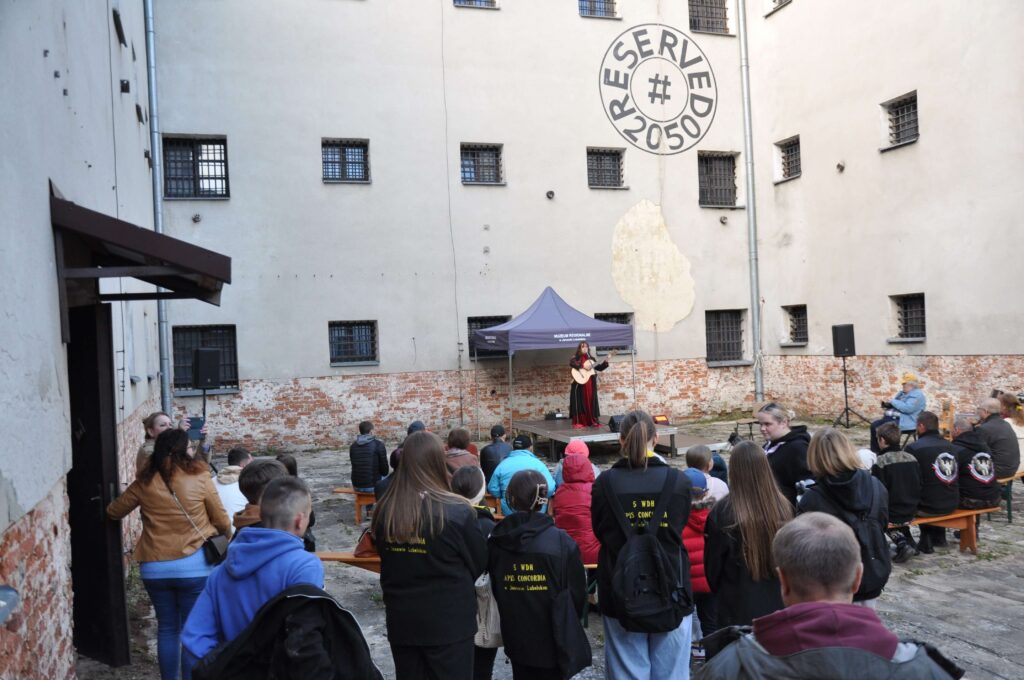 Ludzie siedzący i stojący na dziedzińcu z ceglaną ścianą, widoczna scena z muzykiem grającym na gitarze pod namiotem.