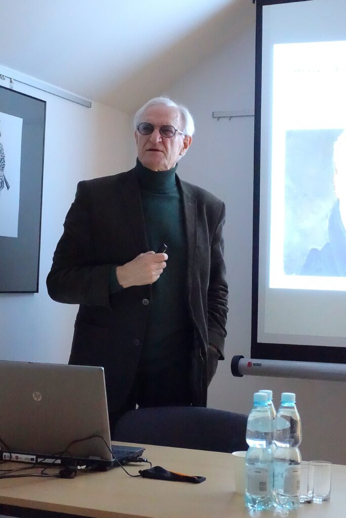 Mężczyzna w okularach i ciemnej marynarce stoi przy stole z laptopem i butelkami wody, na tle jasnego ekranu projekcyjnego.