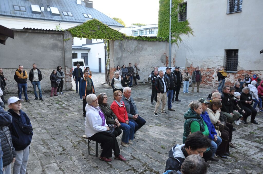 Grupa ludzi siedzących i stojących na brukowanym podwórku otoczonym wysokimi, częściowo pokrytymi zielonym bluszczem ścianami.