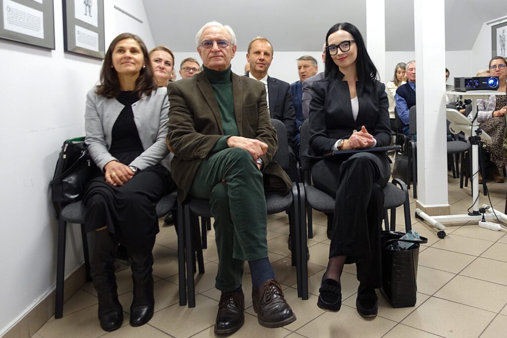 Grupa osób siedzących na krzesłach w jasnym pomieszczeniu, mężczyzna w zielonym garniturze, kobiety w ciemnych i jasnych ubraniach.