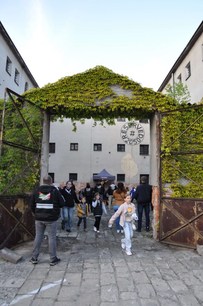 Duża metalowa brama z częściowo porośniętymi zielonym bluszczem kratami, przez którą przechodzi grupa ludzi na brukowanym podwórzu.