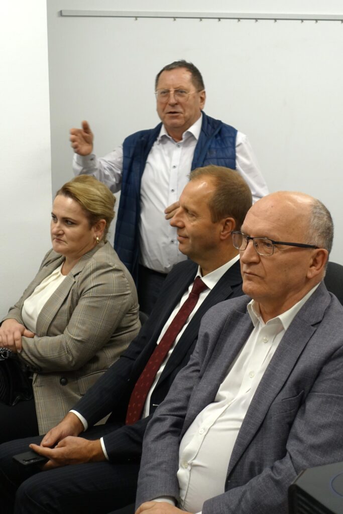 Cztery osoby w formalnych ubraniach siedzą i stoją na tle białej ściany, mężczyzna w kamizelce gestykuluje.