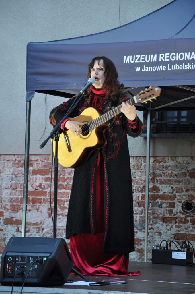 Kobieta w długiej czarnej i czerwonej sukni gra na gitarze klasycznej na scenie z niebieskim namiotem i ścianą z czerwonej cegły.
