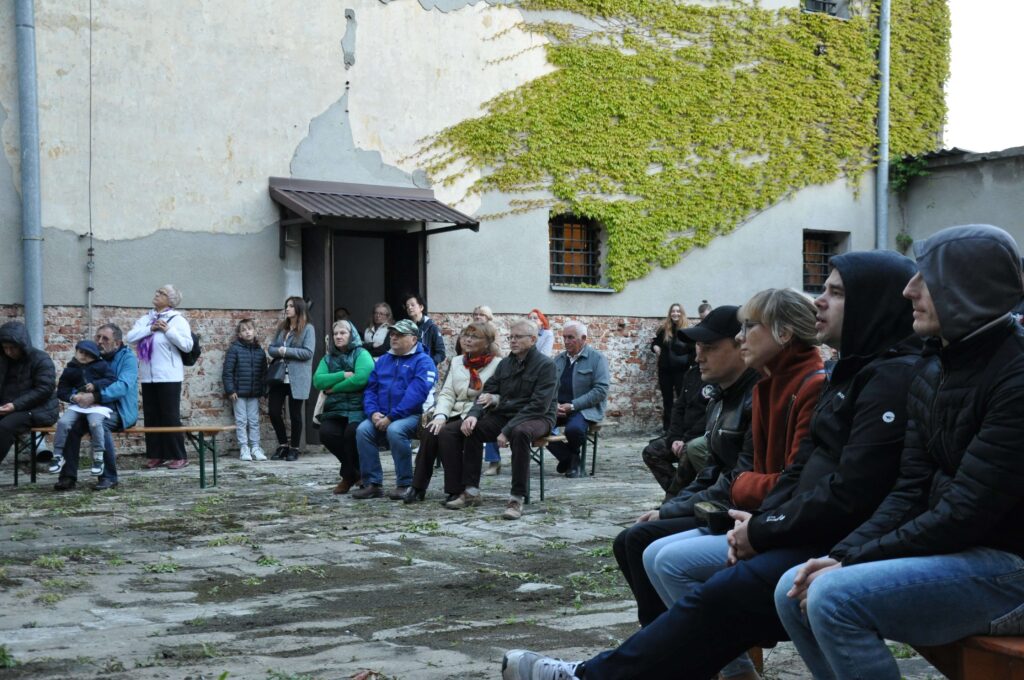 Grupa ludzi siedzących na drewnianych ławkach na kamiennym podwórku przy ścianie z cegły i bluszczem.