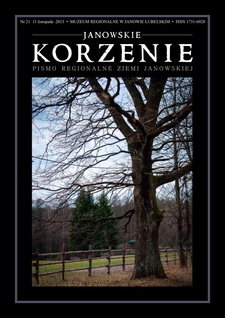 Duże, rozgałęzione drzewo bez liści na tle drewnianego płotu, zielonej trawy i lasu pod niebieskim niebem.