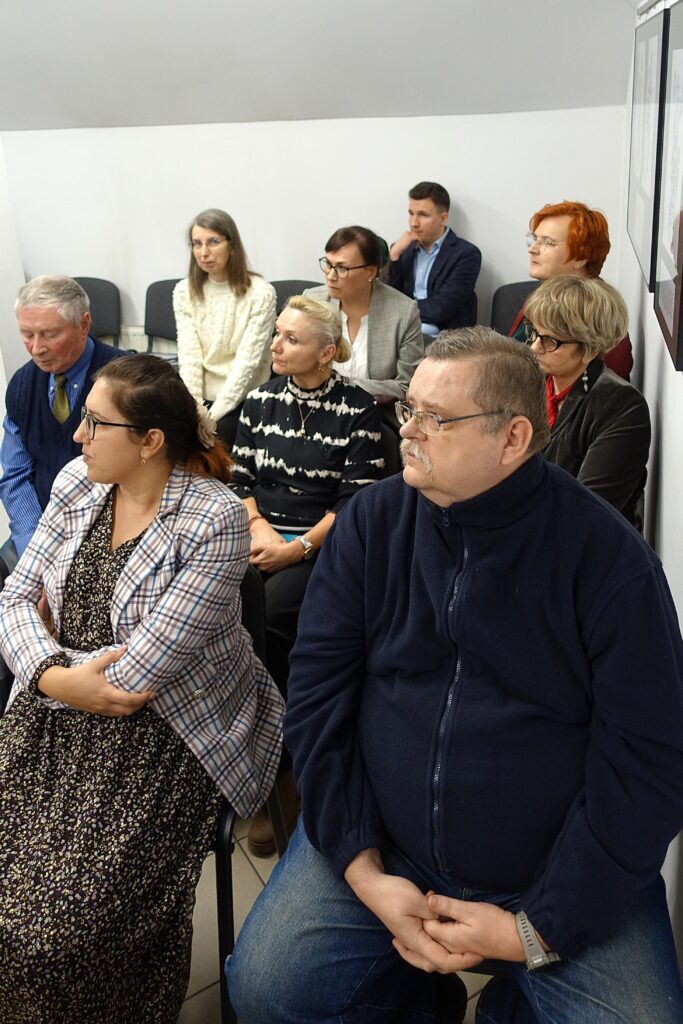 Grupa ośmiu osób siedzących na krzesłach w pomieszczeniu z białymi ścianami, większość w okularach, różne ubrania w stonowanych kolorach.