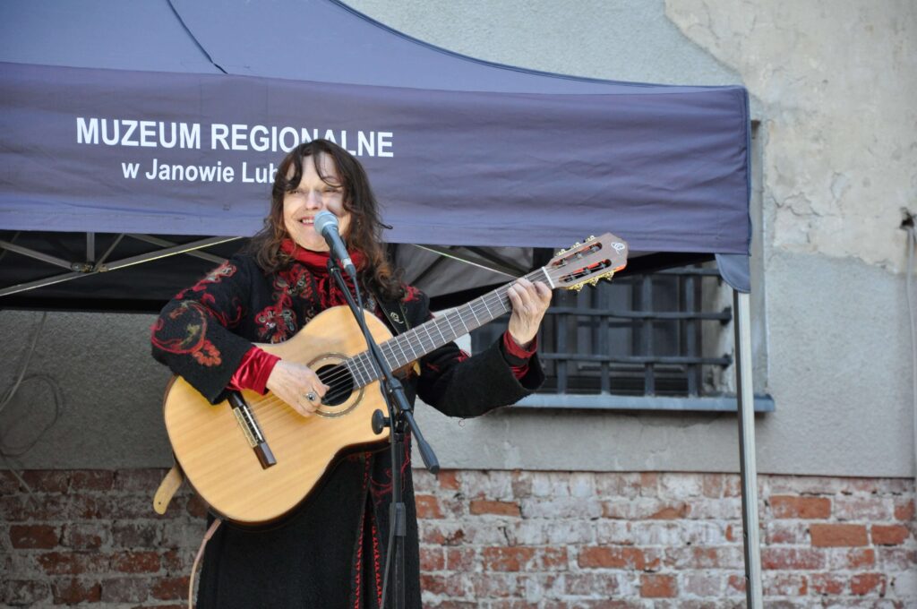 Kobieta w czarnym płaszczu z czerwonymi wzorami gra na gitarze klasycznej przed szarą ścianą i namiotem muzeum.