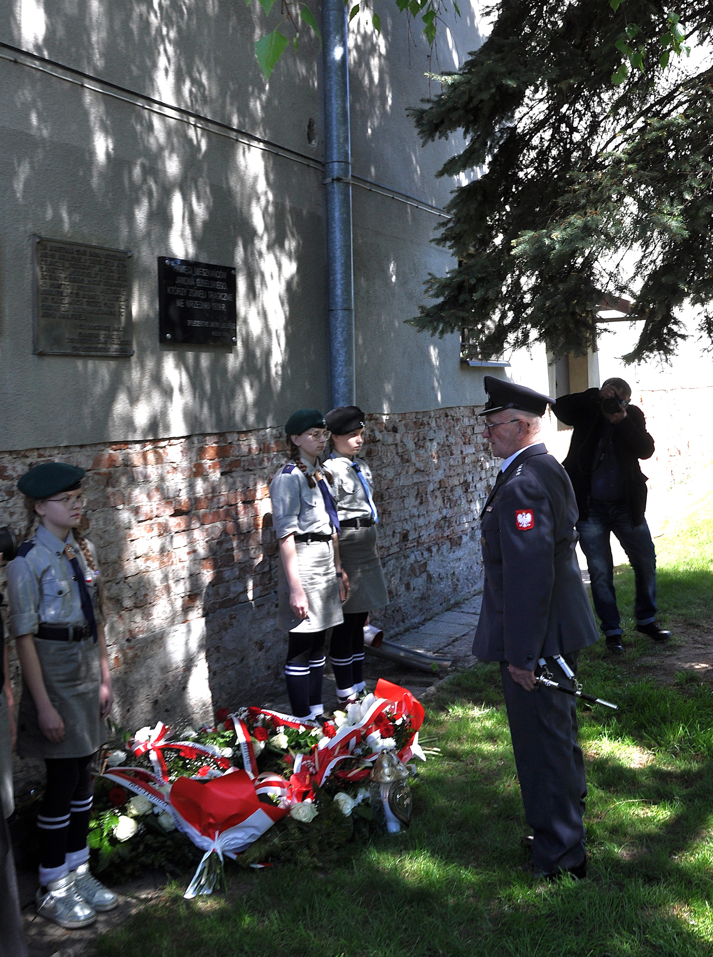 Mężczyzna w mundurze i trzy dziewczynki w harcerskich strojach stoją obok ściany z tablicami i wieńców z biało-czerwonymi wstążkami, fotograf w tle.